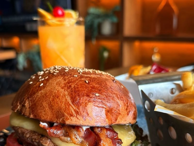 Close-up of a juicy cheeseburger with bacon, crispy fries, and an orange cocktail at Los Canarios, Fiesta Americana Travelty