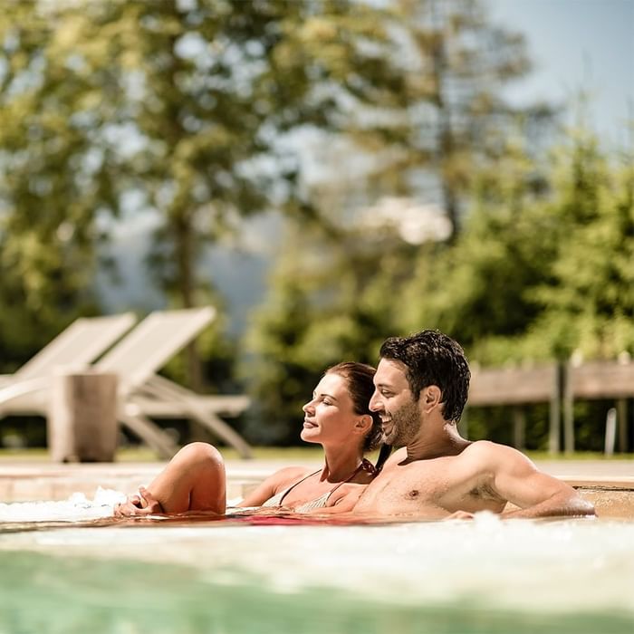 Una coppia si rilassa in una piscina all'aperto circondata da alberi e lettini.