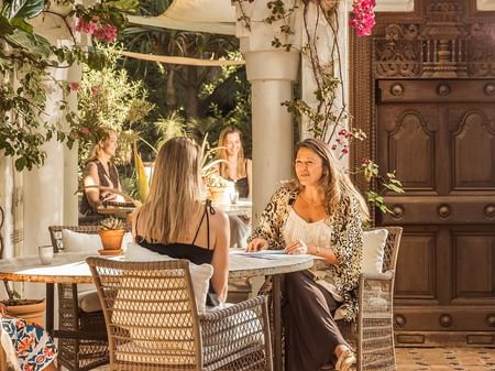 Mujeres conversando en una terraza soleada decorada con flores rosadas y vegetación bajo un toldo de tela natural.