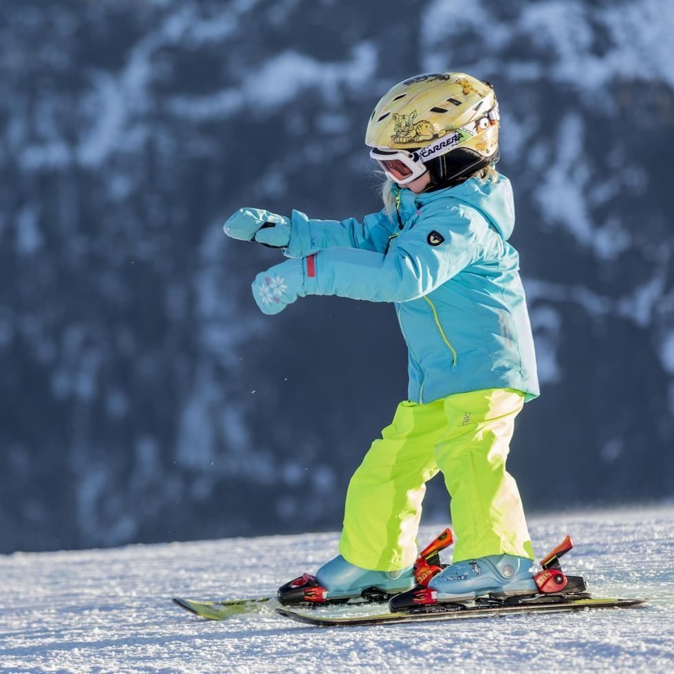 Child skier in blue jacket and yellow pants on snowy mountain slope with snowy mountains in background.
