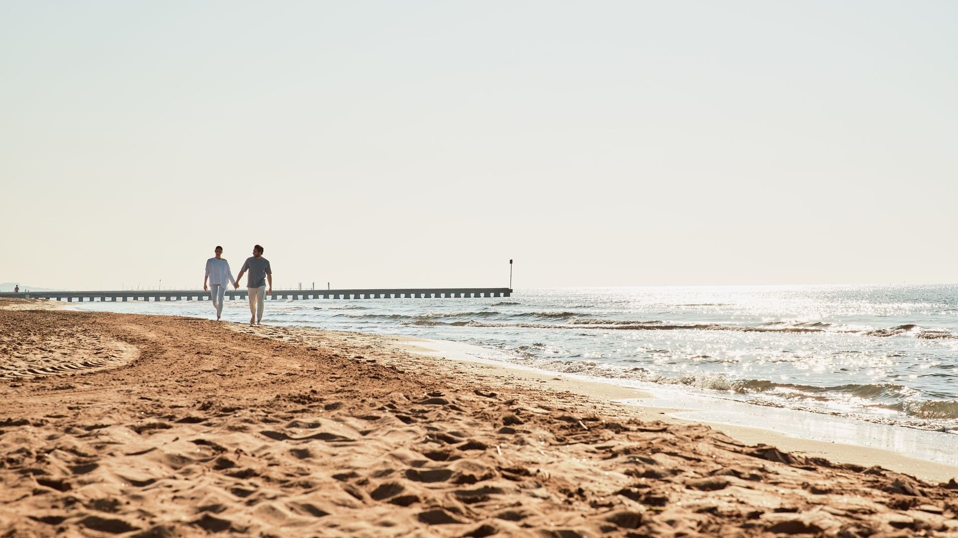 Paar spaziert Hand in Hand am Sandstrand entlang einer Holzsteganlage bei Falkensteiner Group
