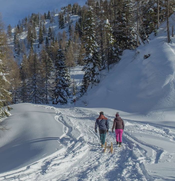 Two people with a sled walking on a snowy trail surrounded by pine trees near Falkensteiner Hotel Sonnenalpe