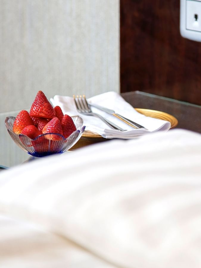 Bowl of strawberries on a bedside table next to a folded napkin with a fork & knife in Superior Room at Warwick Hotel Jeddah
