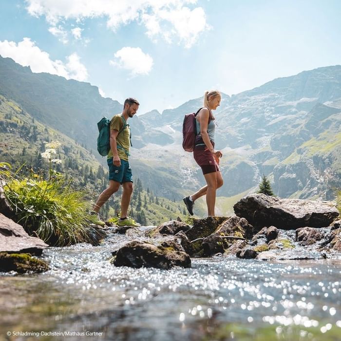 Two hikers with backpacks crossing a rocky stream in a mountainous landscape with blue sky and white clouds.