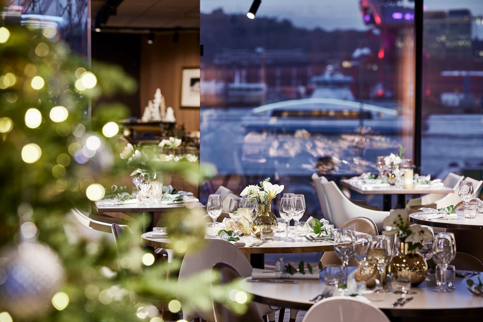 Restaurang med julgran, ställda bord och stora fönster med utsikt över vatten och båtar.