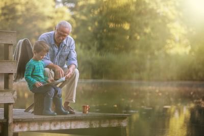 Older man and a young boy sit on a wooden dock by a lake, reading a book together near Fall Creek Marina & Campground