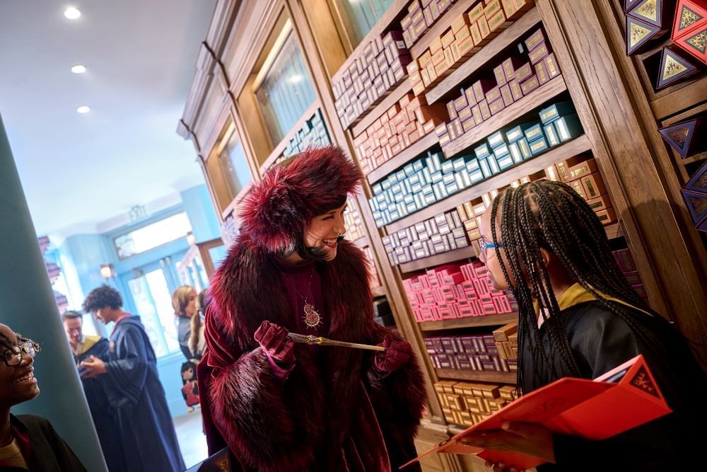 A woman in long, fur-lined magenta robes and a coordinating hat shows a wand to a young girl in wizarding robes.