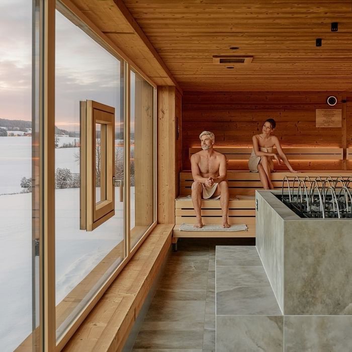 Sauna moderna con vista su paesaggio innevato, due persone rilassate su panca in legno.