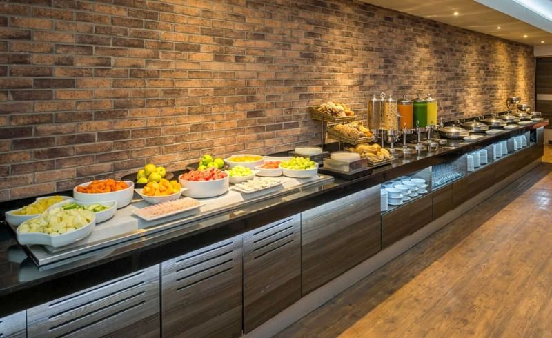 Buffet de desayuno con frutas, jugos y pasteles frente a pared de ladrillo en Real Inn Perinorte