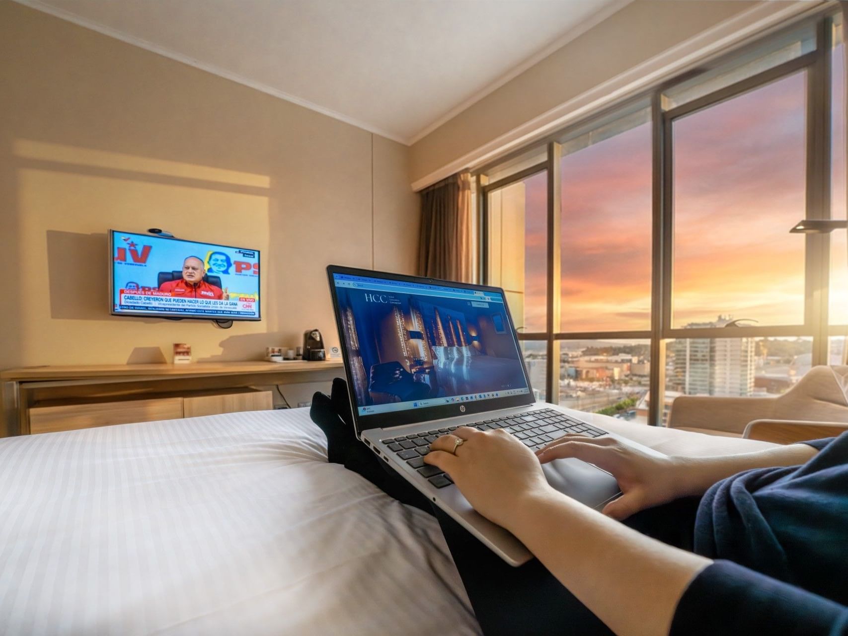 Una persona utilizando un ordenador portátil en una cama con vistas a la ciudad en el hotel HCC Costanera de Concepción.