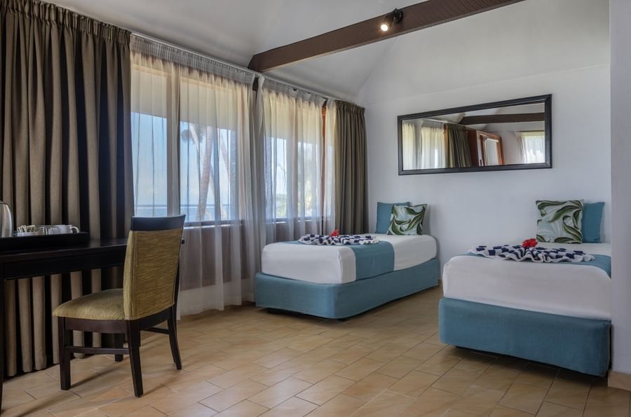 Twin beds with teal bases by a large window & wall Mirror in Beach Front Bure with Plunge Pools at Tambua Sands Beach Resort