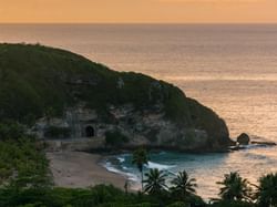 Dark tunnel entrance in a rocky, green cliff face above a tropical beach at sunset in Tunel de Guajataca near Royal Isabela