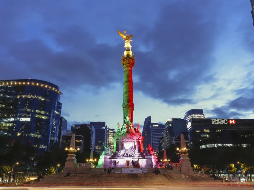 Ángel de la Independencia iluminado con colores nacionales al anochecer cerca de Camino Real Hotels