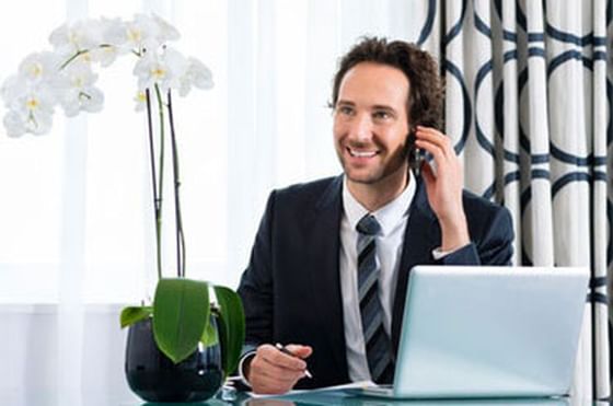 Man in a suit talking on a phone beside his laptop and an orchid