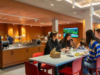 Guests enjoying breakfast together at a communal table in a modern dining area with warm orange walls and buffet stations