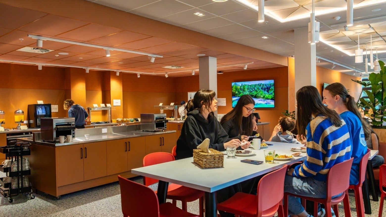 Guests enjoying breakfast together at a communal table in a modern dining area with warm orange walls and buffet stations