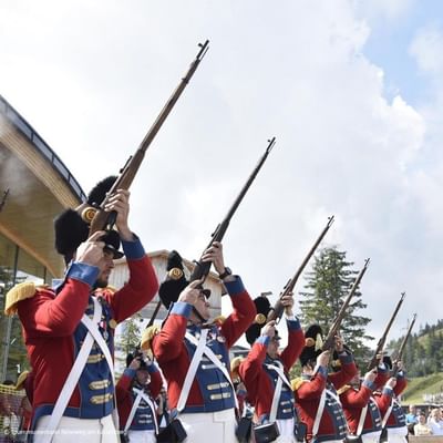 Soldiers in red and white uniforms hold rifles in salute position, standing in formation.