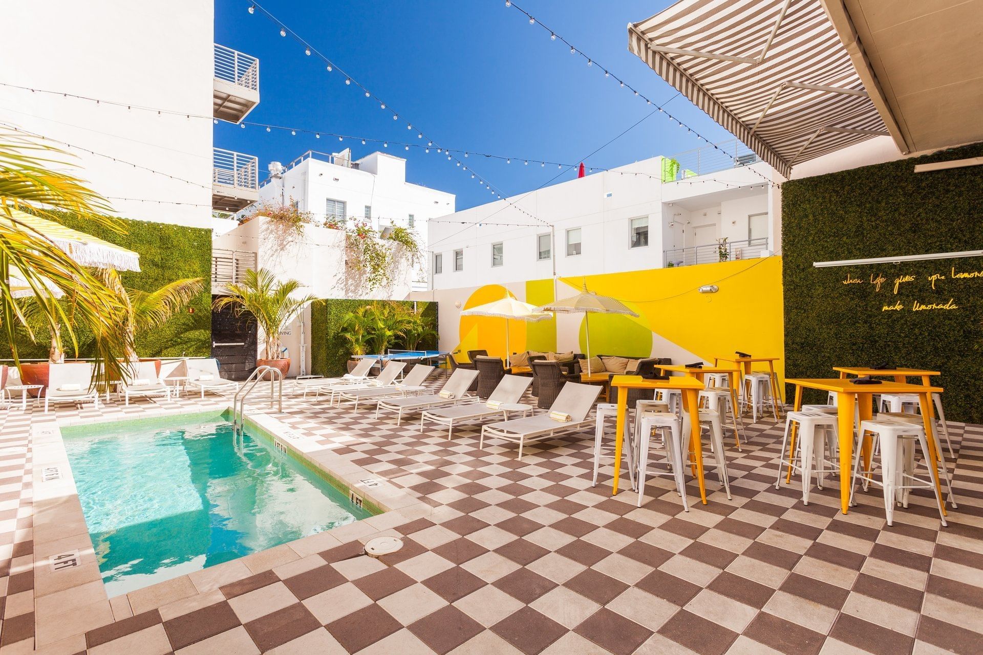 A poolside area with lounge chairs, tables, and umbrellas, surrounded by greenery and a yellow wall.