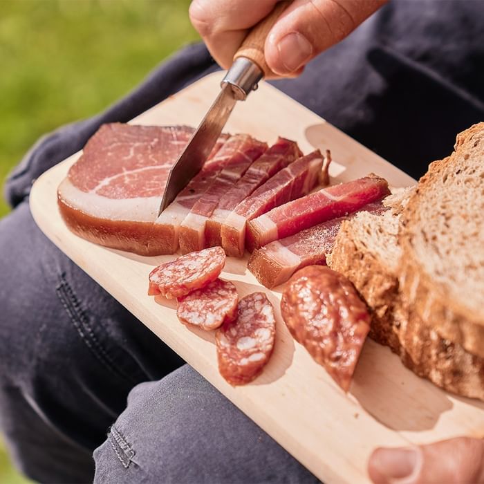 Person slicing prosciutto and salami on a wooden board with a knife at Falkensteiner Hotel Montafon