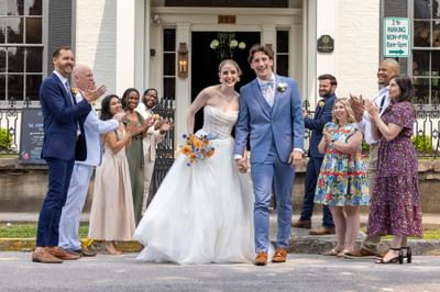 Newlywed couple holding hands while guests applaud outside the white brick entrance at The Ashbrook Hotel