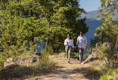 Two people walk on a trail by a lake, surrounded by trees and rocks.