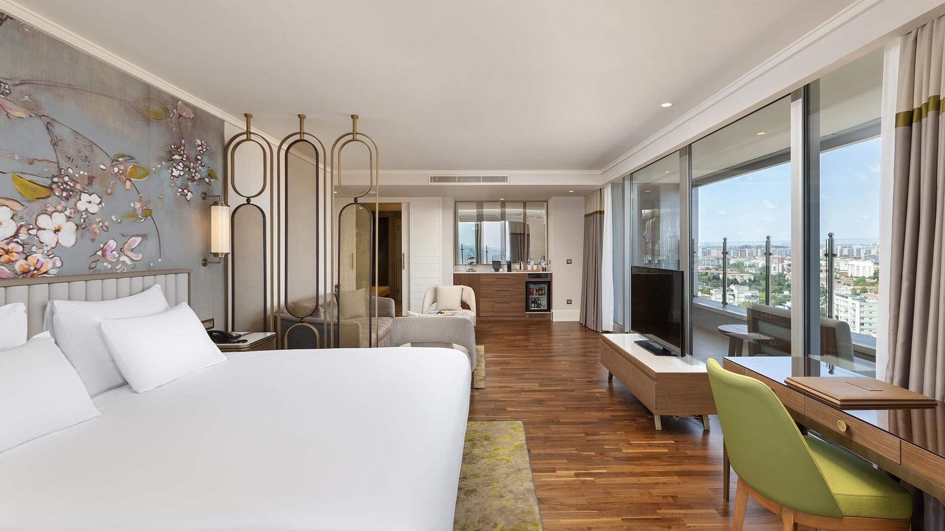 A luxurious hotel room with a large bed, floral wall art, a desk and chair, and a panoramic city view through floor-to-ceiling windows in Junior Suite at Titanic Port Bakırköy