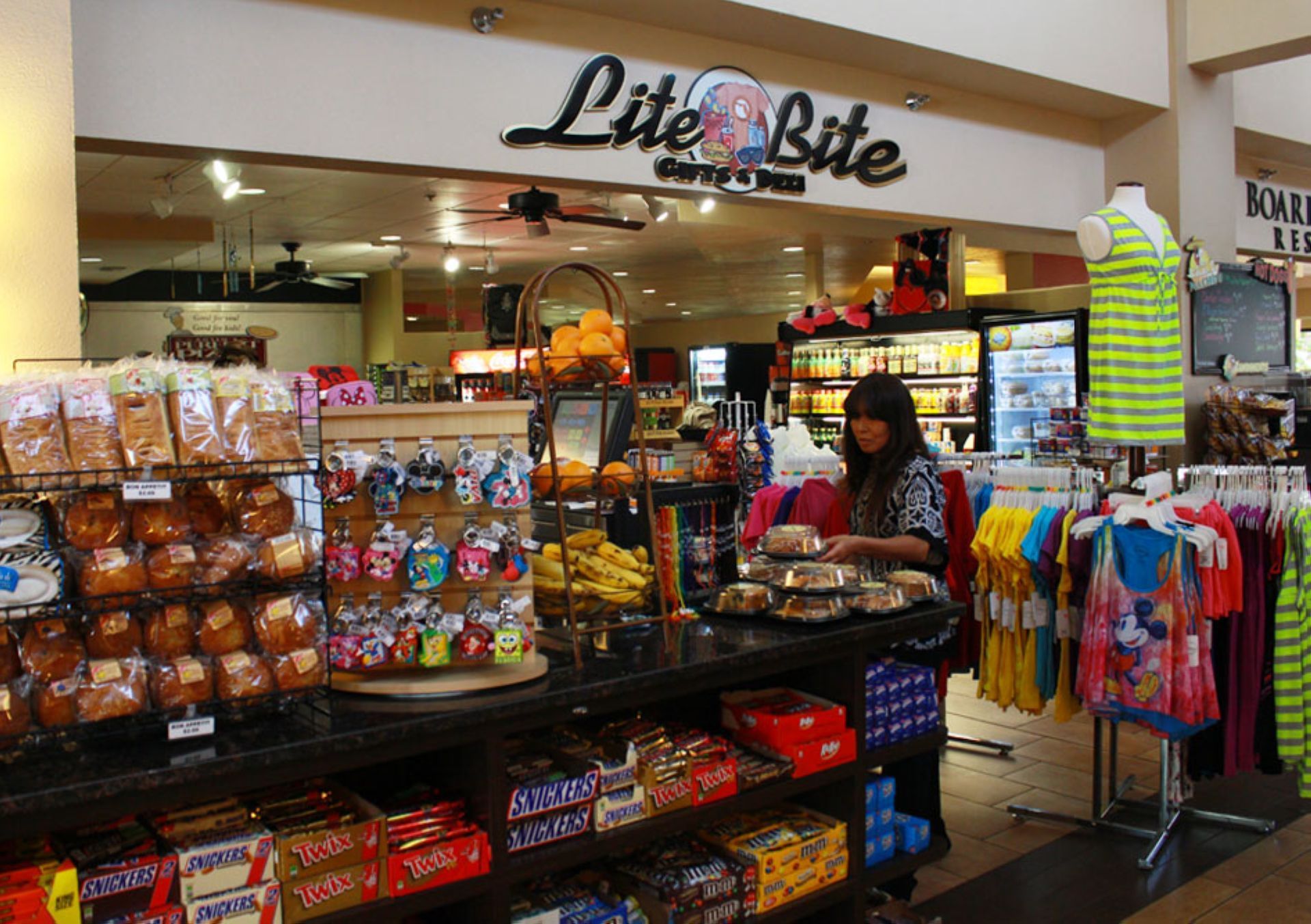 A convenience store with a counter displaying various snacks and drinks, as well as clothing racks and a refrigerator.