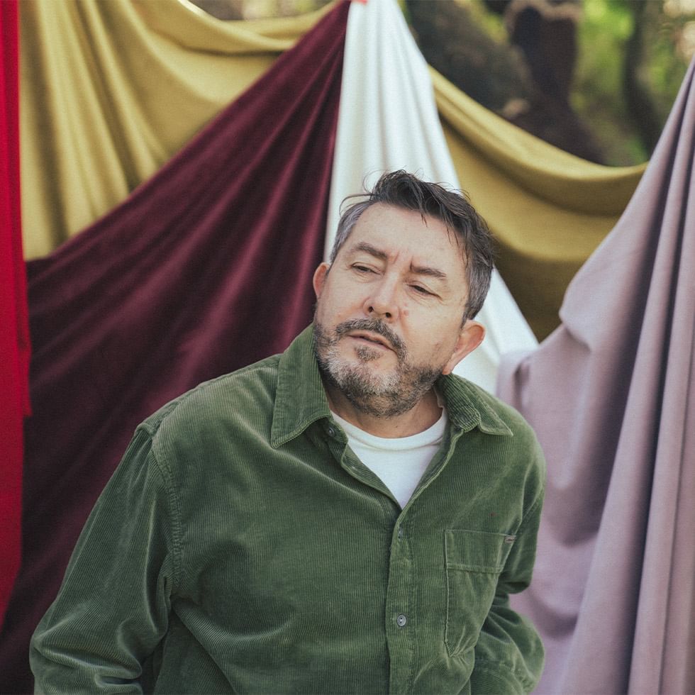 Man with closed eyes and beard in green shirt stands before a backdrop of multicolored fabrics.