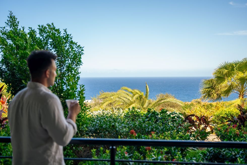 Persoon geniet van een drankje op balkon terwijl hij uitkijkt op de oceaan in Deluxe Ocean View Room bij Golden Rock Resort