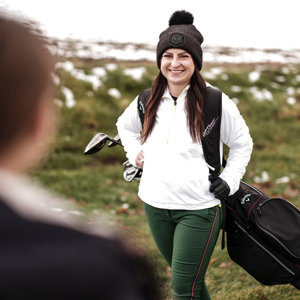 Smiling woman holding a golf bag and clubs at a golf course near Falkensteiner Balance Resort Stegersbach