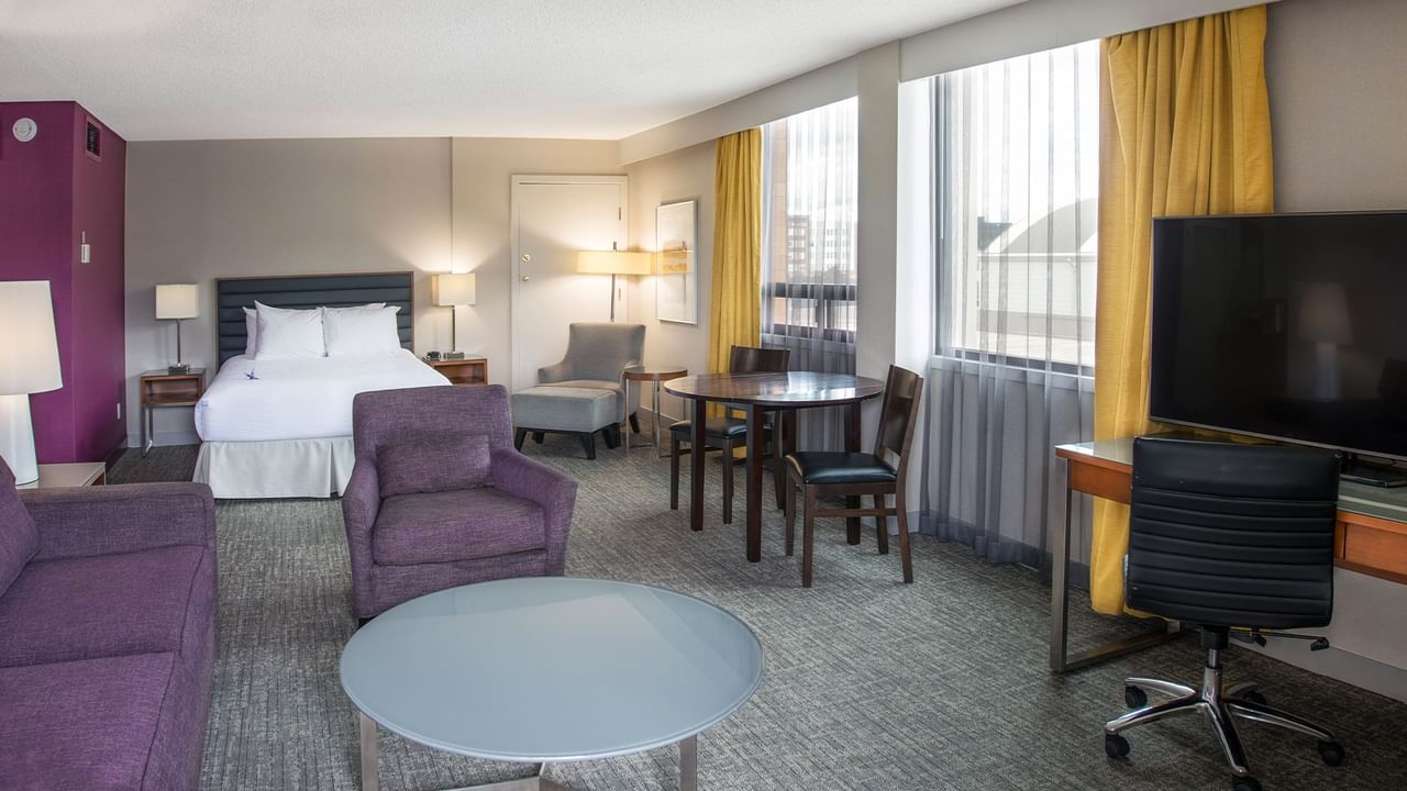 A hotel suite with a bed, couch, chairs, table, TV, and large windows.