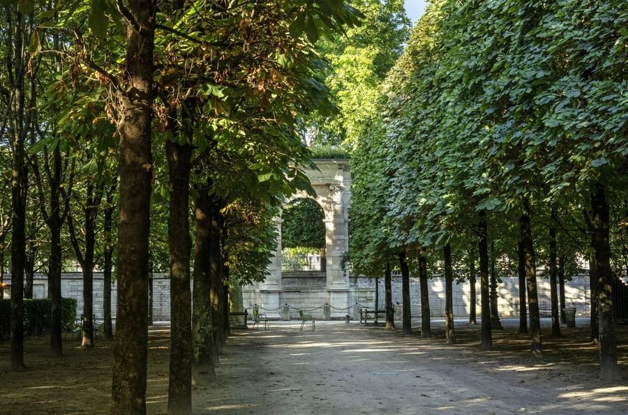 Stone archway placed under a dense canopy of lush green trees by a walking path near Hotel Westminster Paris