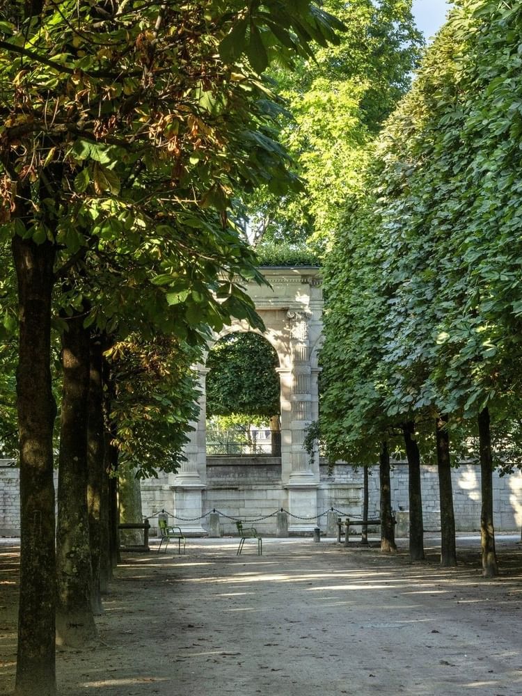 Stone archway placed under a dense canopy of lush green trees by a walking path near Hotel Westminster Paris