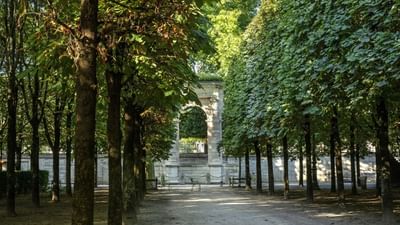 Stone archway placed under a dense canopy of lush green trees by a walking path near Hotel Westminster Paris