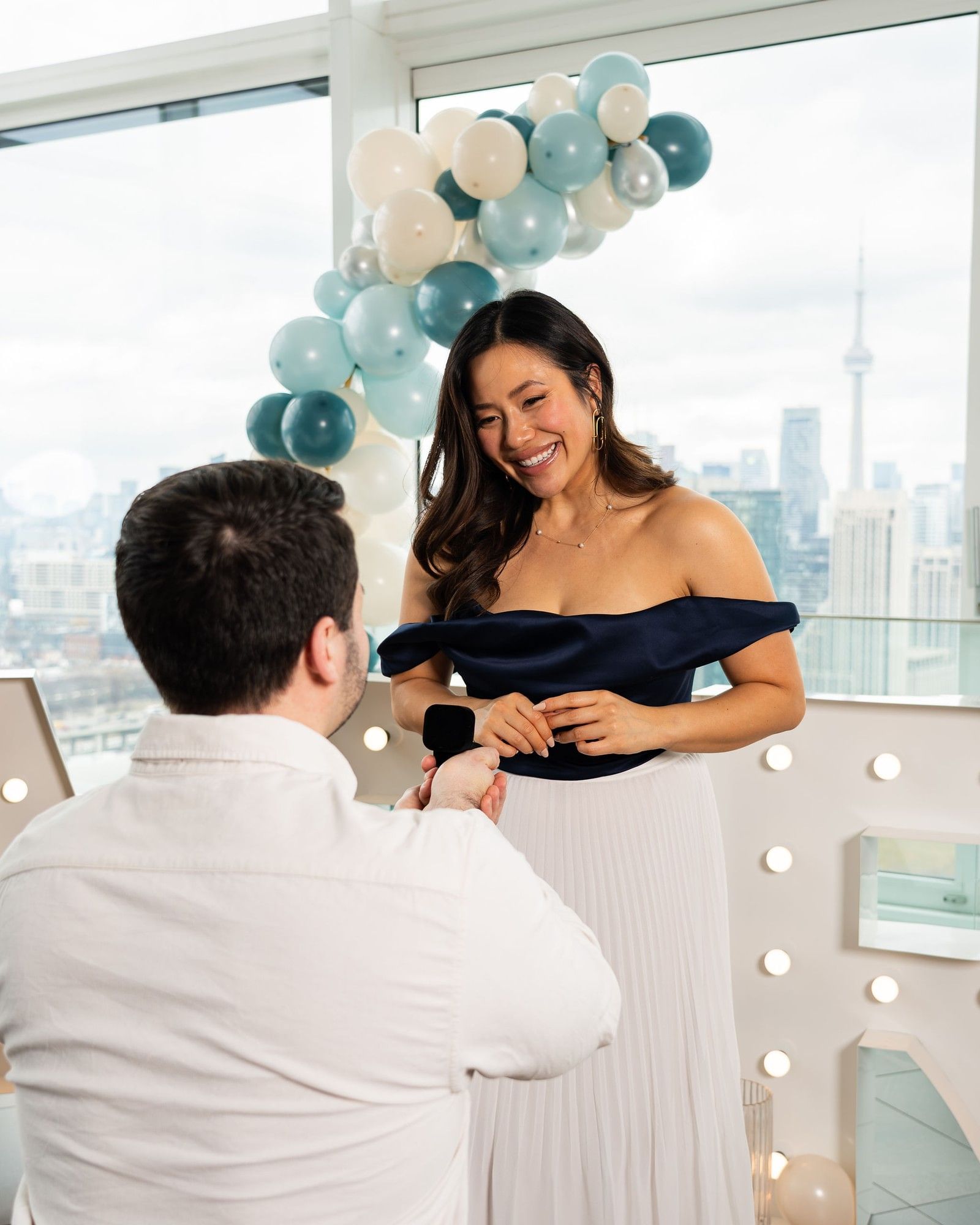 Man proposing with a ring by blue and white balloons under bright windows overlooking the CN Tower at Hotel X Toronto