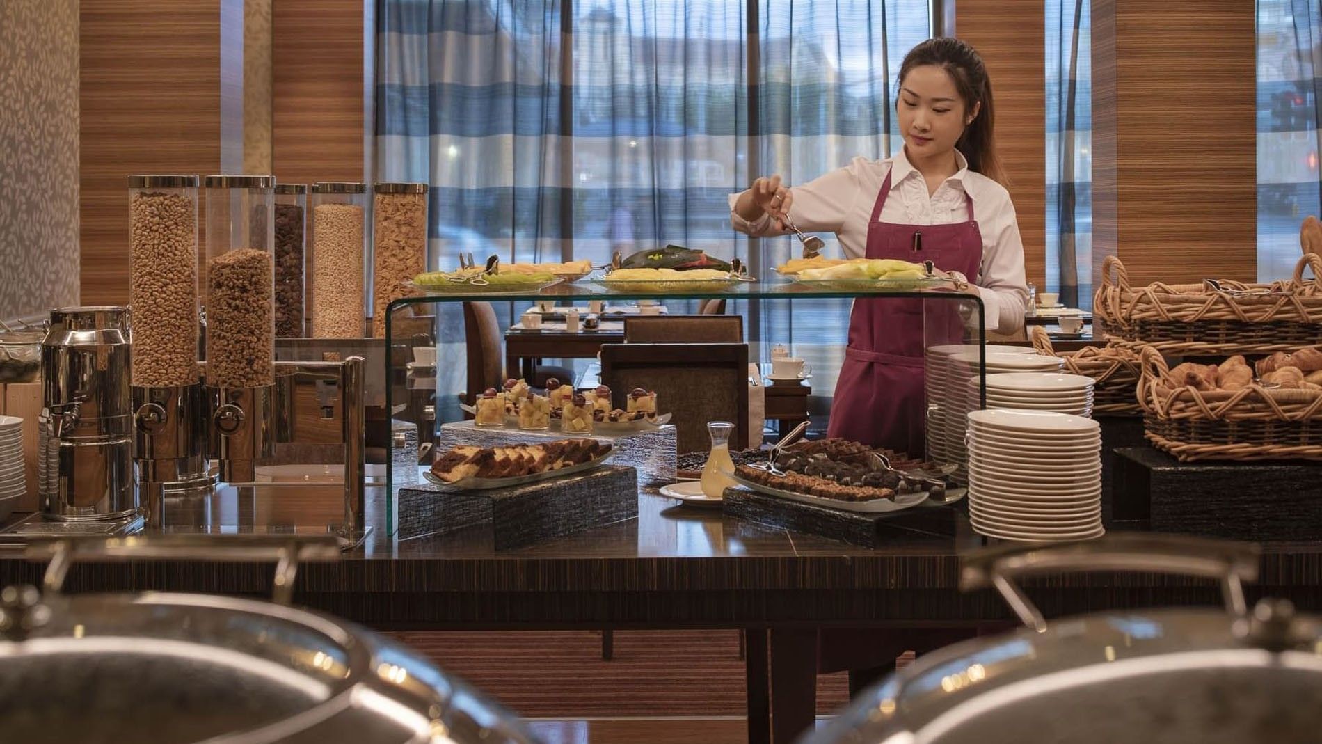 Attendant serving fresh fruit by cereal dispensers under a glass shelf in Breakfast at Warwick Geneva