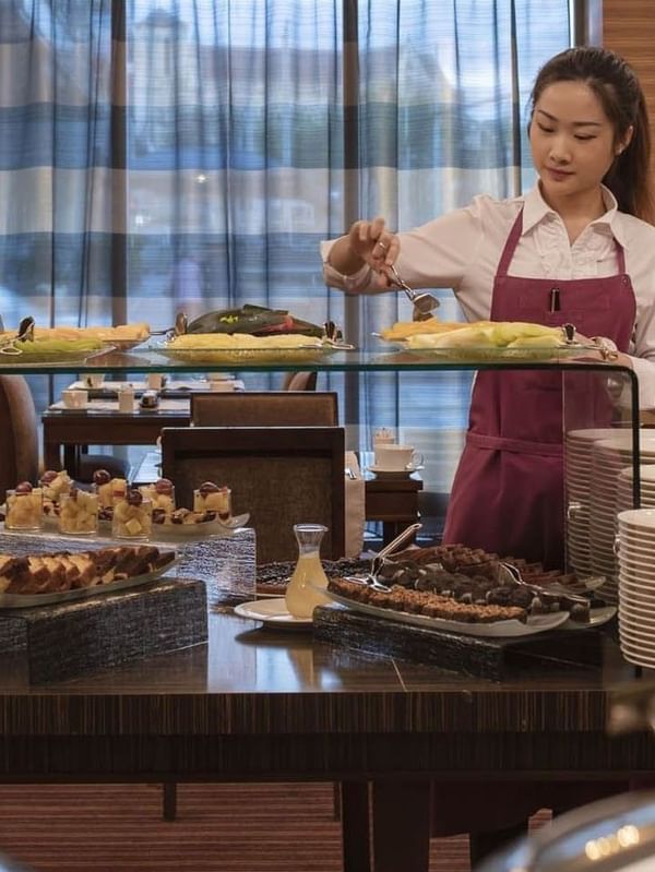 Attendant serving fresh fruit by cereal dispensers under a glass shelf in Breakfast at Warwick Geneva
