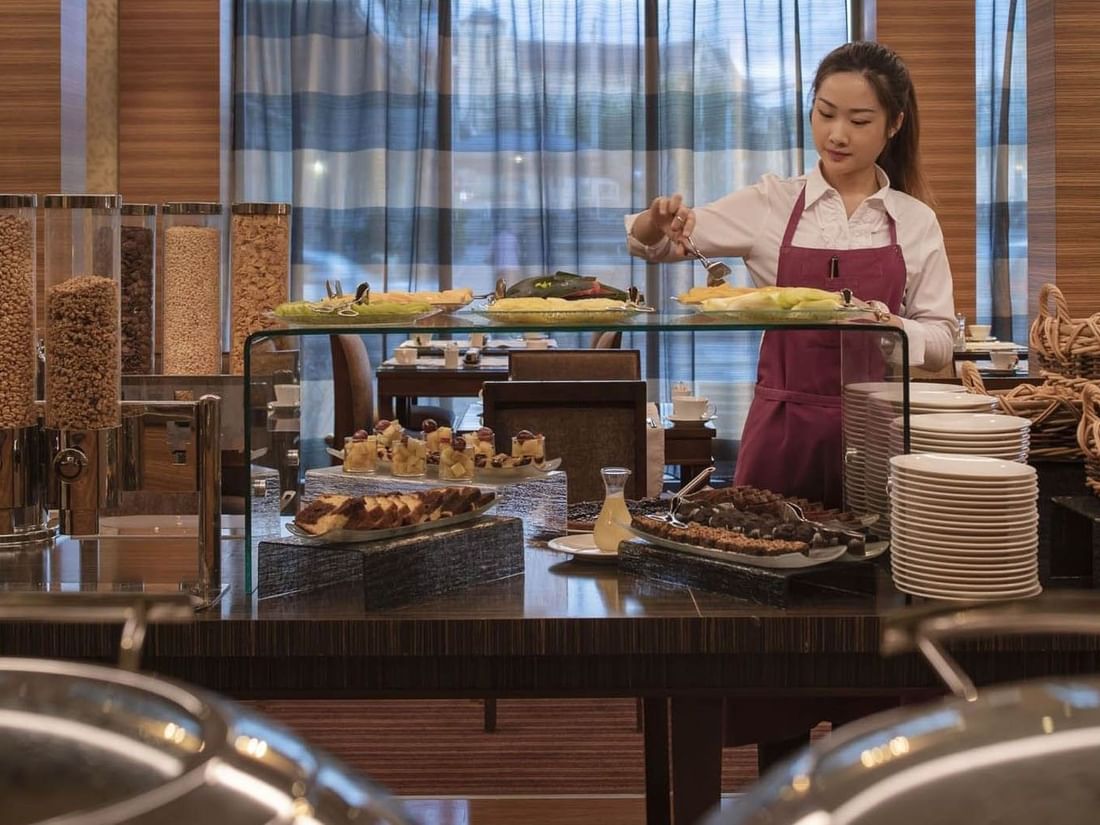 Attendant serving fresh fruit by cereal dispensers under a glass shelf in Breakfast at Warwick Geneva