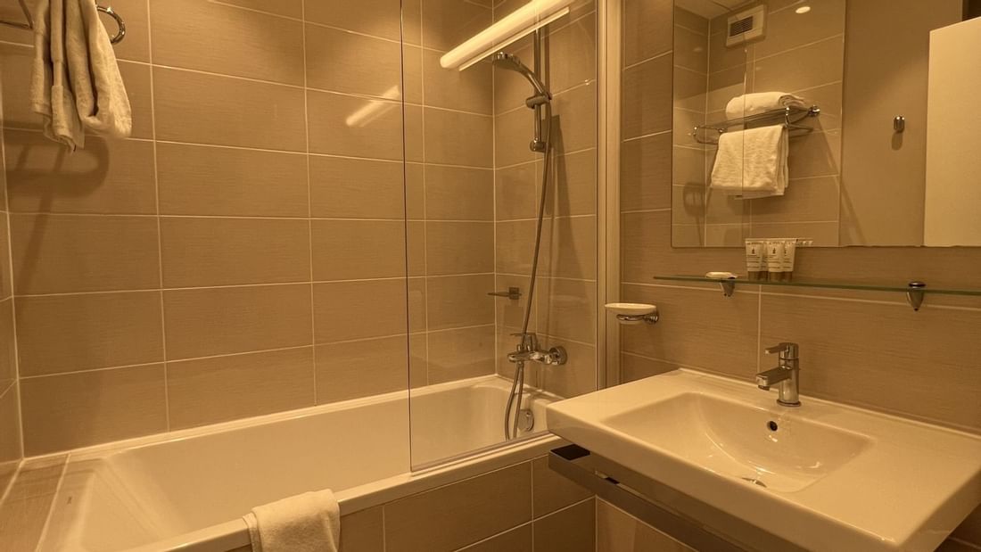 Salle de bain avec baignoire, lavabo mural, miroir lumineux et carrelage clair.