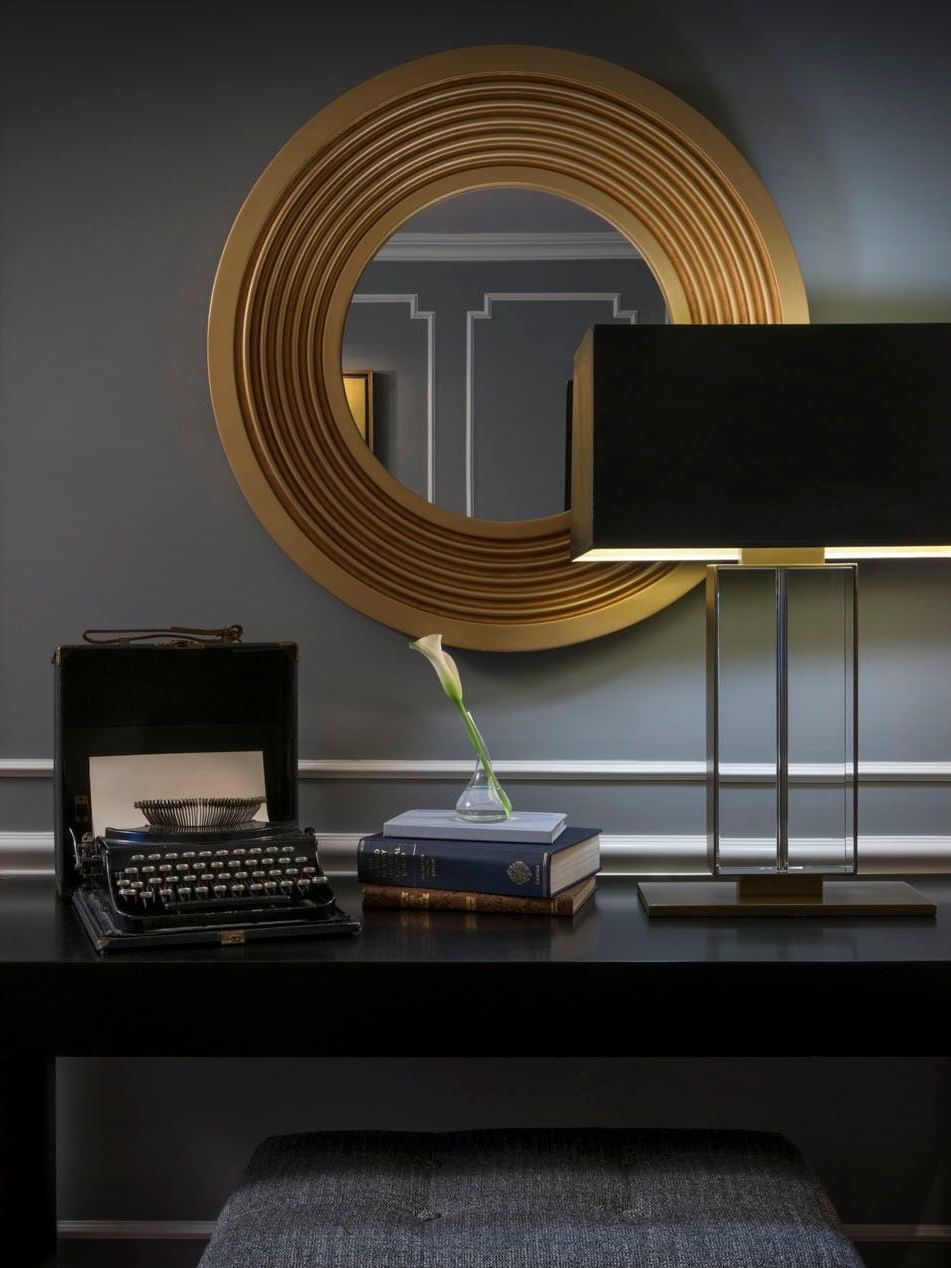 Black typewriter and books sit on dark desk beneath a large and round gold wall mirror in Randolph Suite at Warwick New York