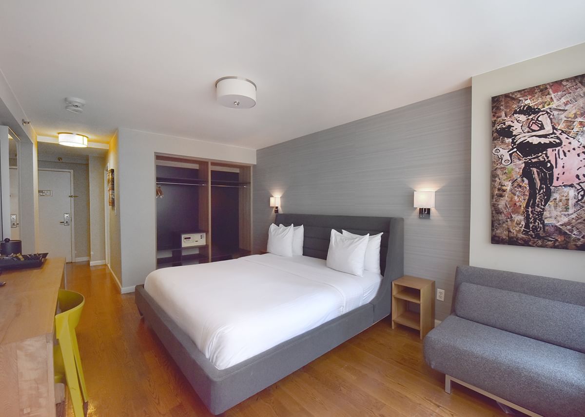 A hotel room with a king-sized bed, gray upholstered headboard, and a gray sofa against a gray wall with a large piece of artwork.