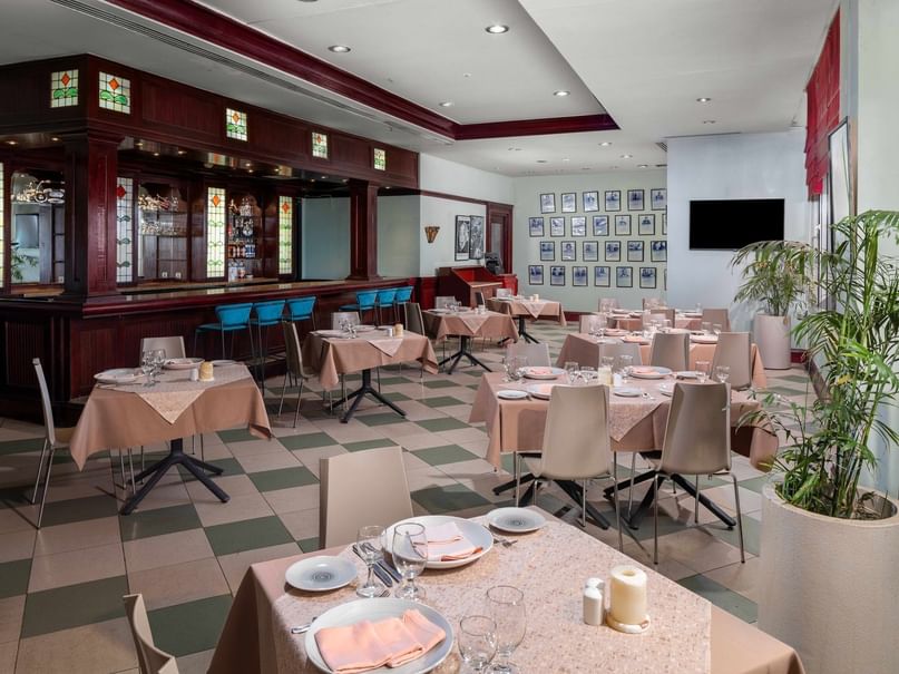 Elegant restaurant with checkered floor, set tables, bar area, framed pictures, and a large potted plant.