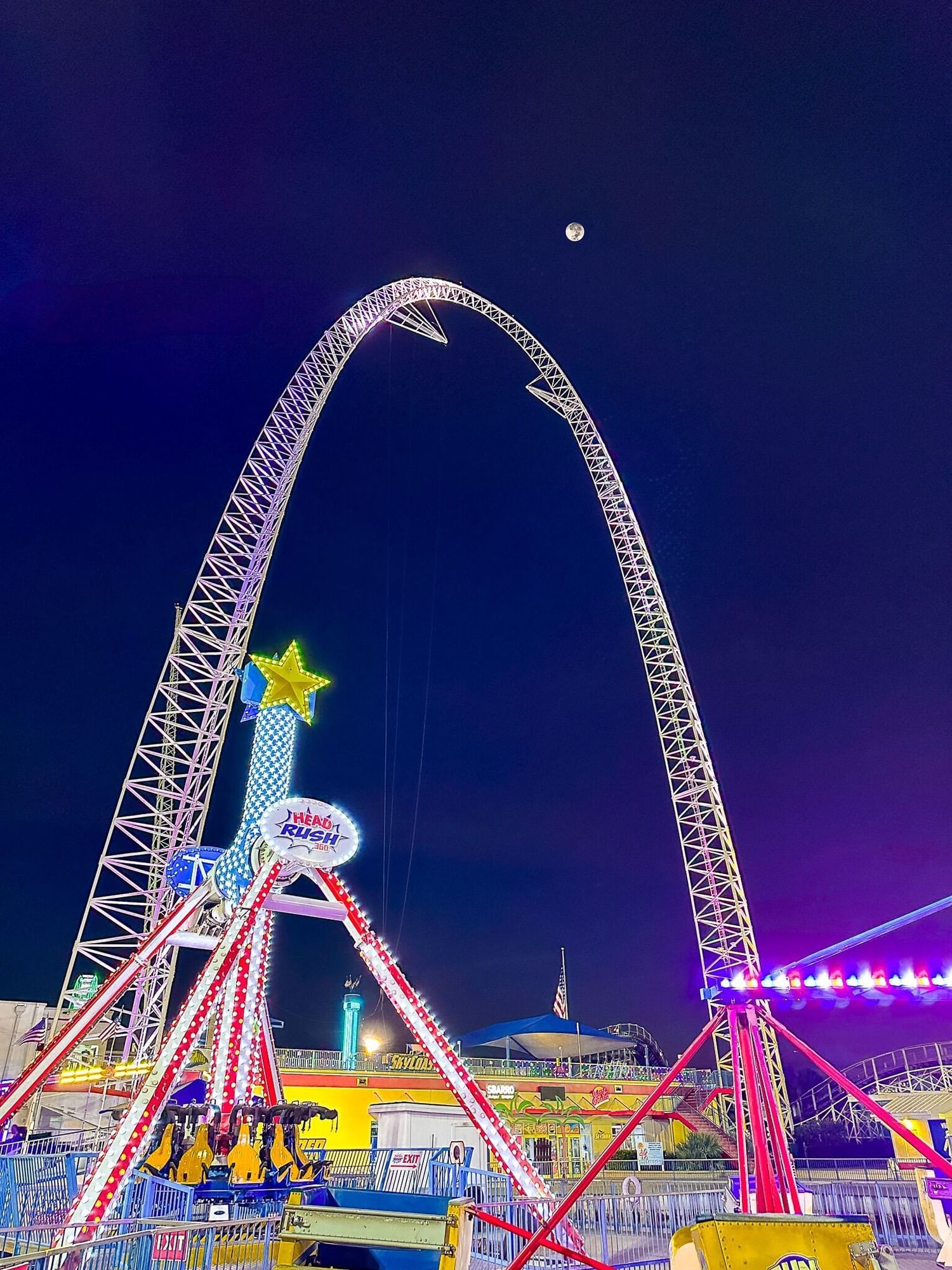 At night, a large glowing archway surrounded by carnival rides reaches into the sky with the moon glowing in the distance.