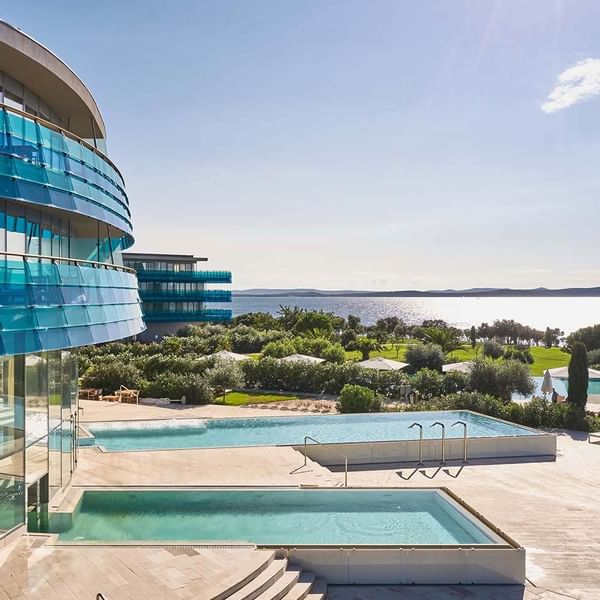 Modernes Hotelgebäude mit zwei Pools, umgeben von Grün und mit Blick auf das Meer.