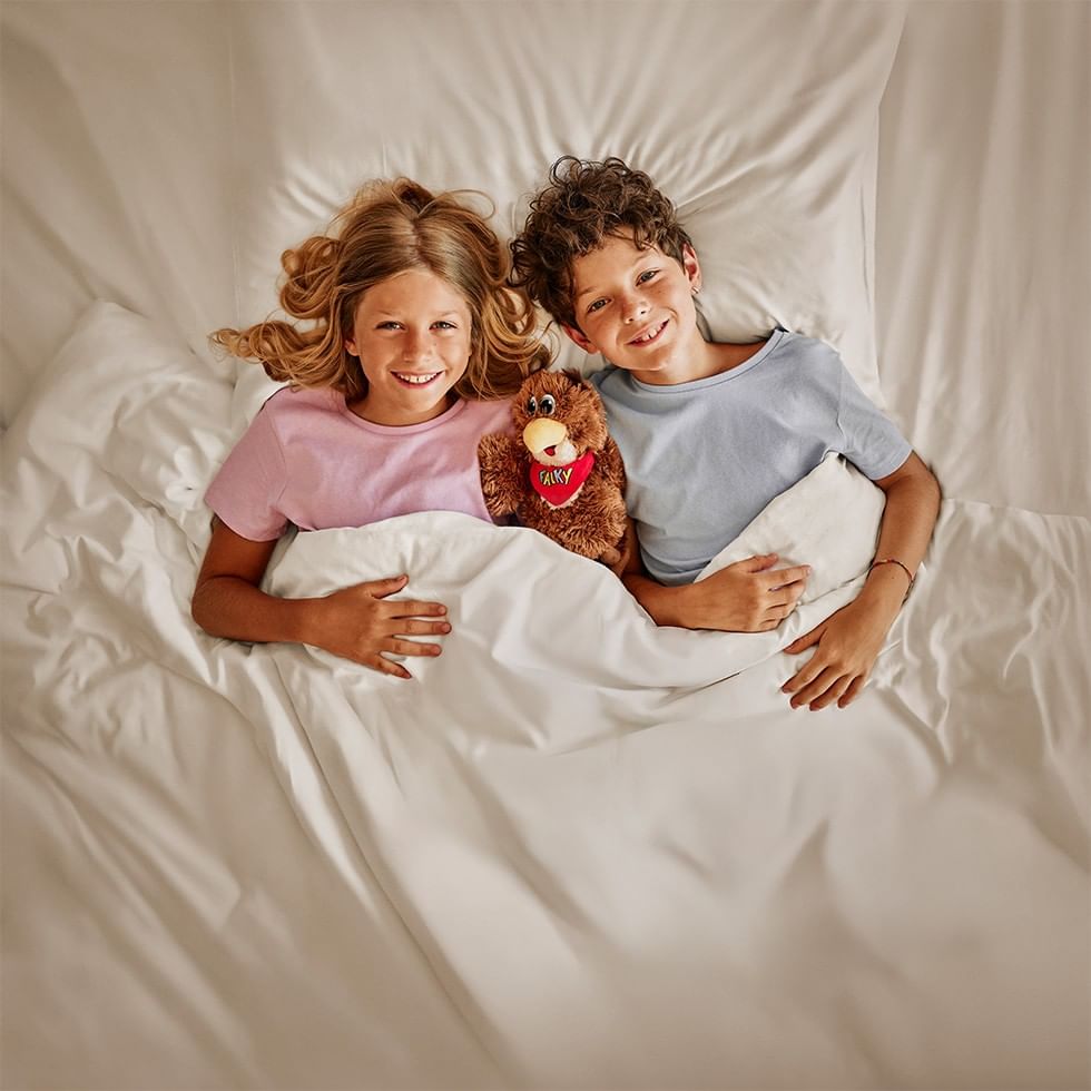 Due bambini sorridenti con un peluche, sotto una coperta bianca, in un letto matrimoniale.