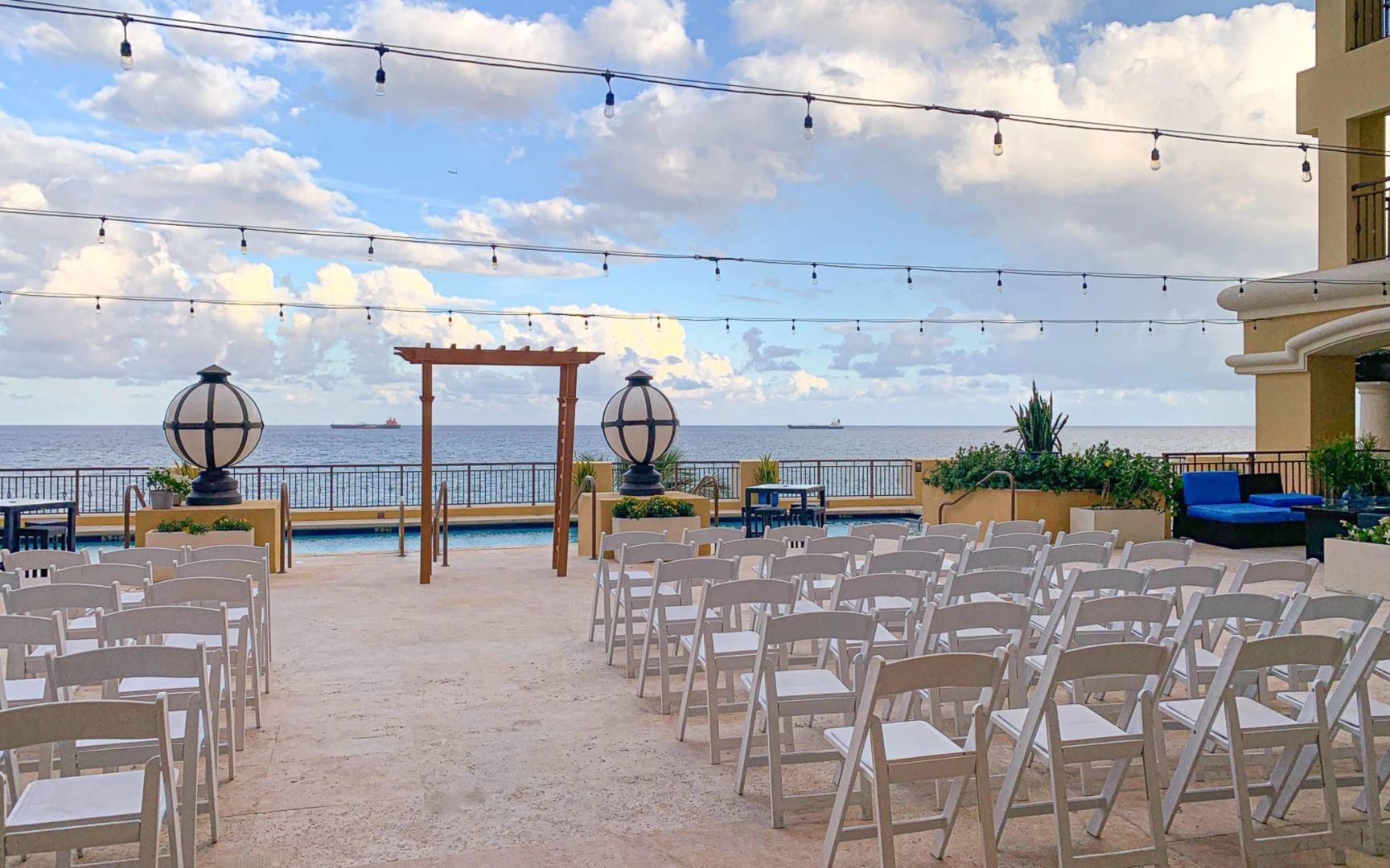 An outdoor wedding setup with white chairs, a pergola, and string lights overlooking the ocean.