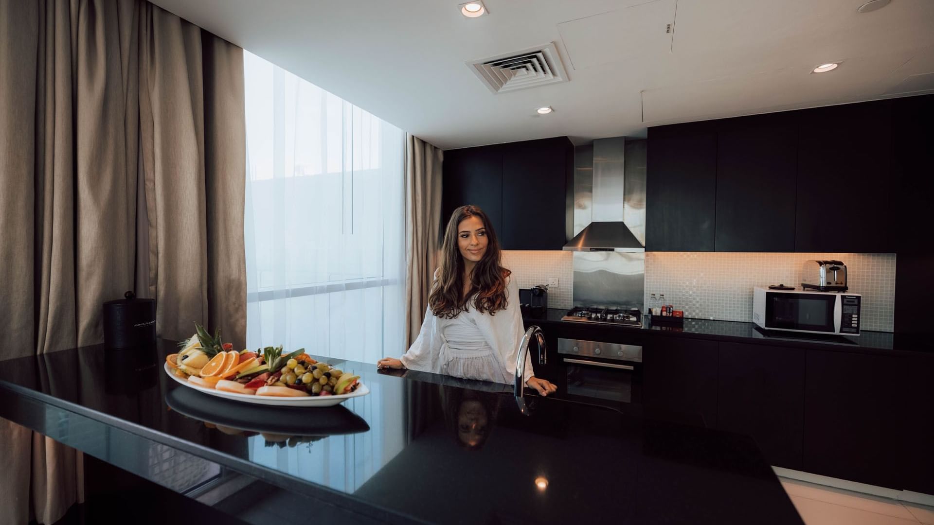 A lady in a white dress stands in the kitchenette in a hotel room at DAMAC Maison Distinction