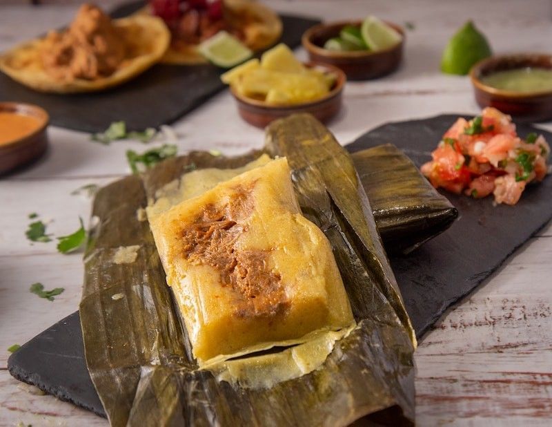Tamale in a banana leaf on a black slate plate, with various other Latin American dishes served near Real Inn Nuevo Laredo