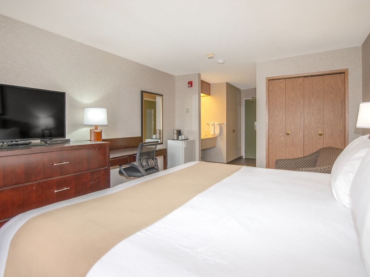 A hotel room with a bed, dresser, TV, chair, and a small refrigerator.