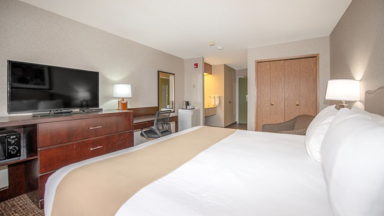 A hotel room with a bed, dresser, TV, chair, and a small refrigerator.